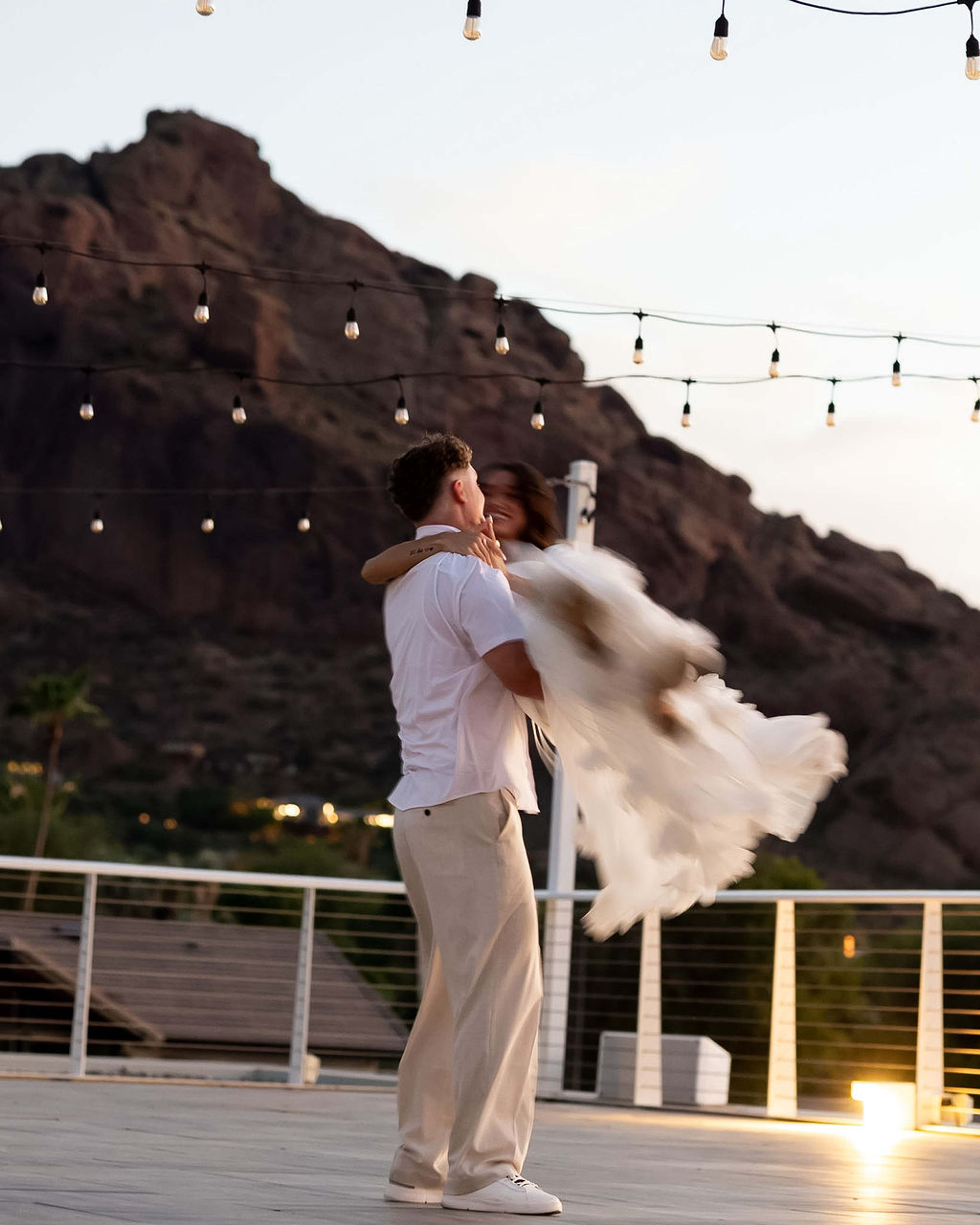 A man lifts and spins a woman on a terrace, her skirt flaring out, Camelback Mountain and string lights behind