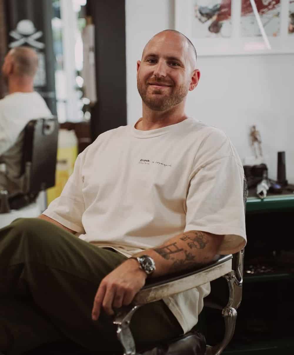 Tommy sitting sideways across a chrome barber chair in a white tee and olive trousers, with a closely shaved head and a short beard.