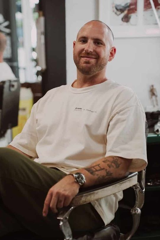 Tommy sitting sideways across a chrome barber chair in a white tee and olive trousers, with a closely shaved head and a short beard.
