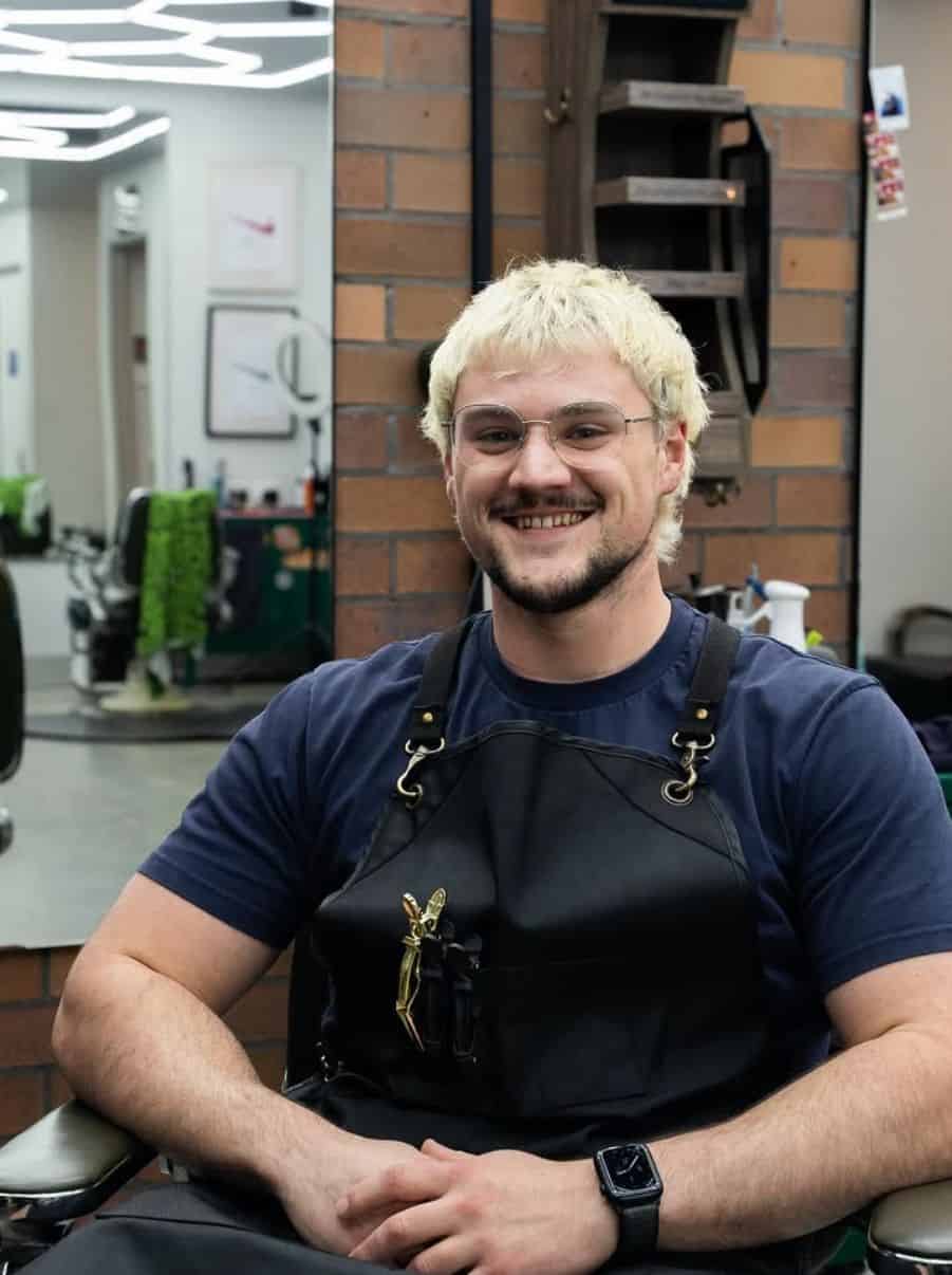 Callum grinning in a barber chair in a navy tee and a black cutting apron, with bleached blonde hair and clear-framed glasses.