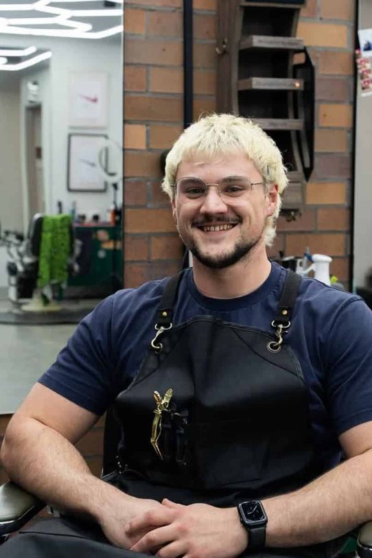 Callum grinning in a barber chair in a navy tee and a black cutting apron, with bleached blonde hair and clear-framed glasses.