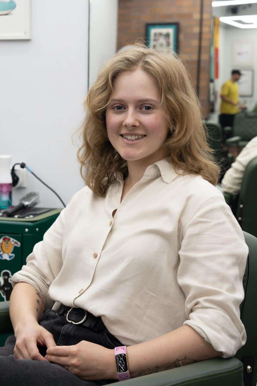 Georgina sitting in a barber chair in a cream linen shirt, with shoulder-length wavy blonde hair and a small tattoo on one forearm.