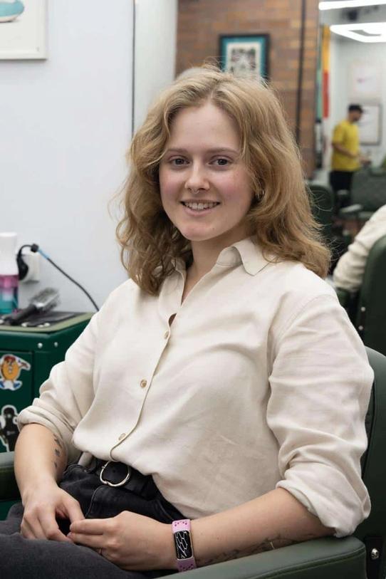 Georgina sitting in a barber chair in a cream linen shirt, with shoulder-length wavy blonde hair and a small tattoo on one forearm.