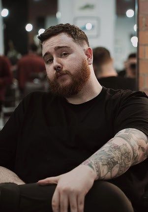 Conor leaning back in a barber chair in a black tee, with a full ginger beard, short dark hair and a heavily tattooed forearm.