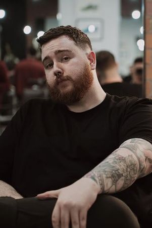 Conor leaning back in a barber chair in a black tee, with a full ginger beard, short dark hair and a heavily tattooed forearm.