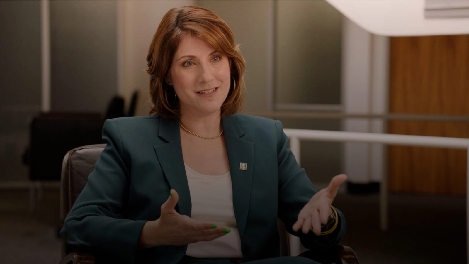 A smiling woman with reddish-brown hair in a teal suit jacket gestures with both hands in a professional setting.