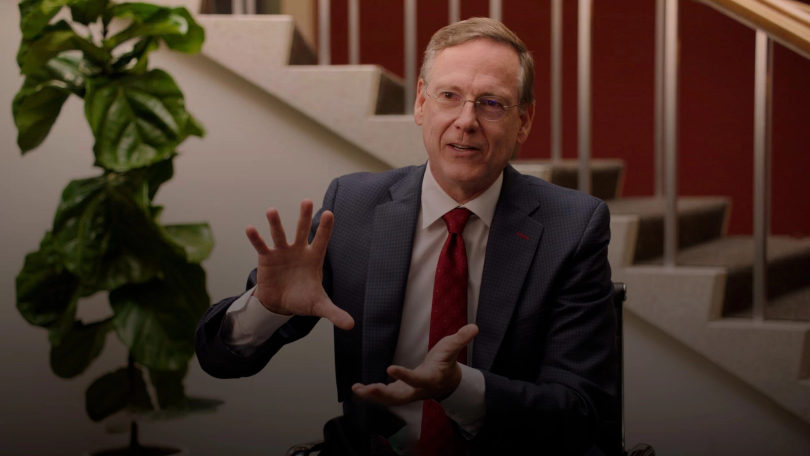 A man in a suit and glasses speaks and gestures, with a large plant and staircase behind him.