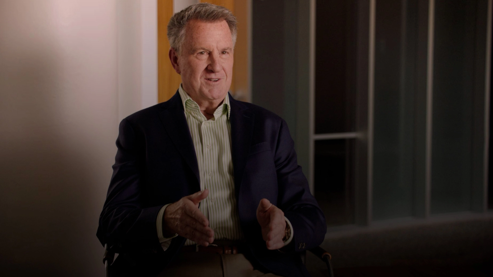 A man in a suit jacket and striped shirt speaks and gestures with his hands in an indoor setting.