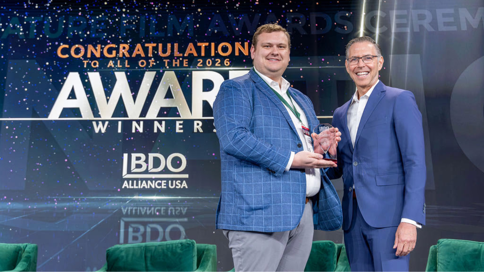 Two smiling men on stage; one in a blue plaid jacket accepts a clear award from another in a blue suit, with "AWARD WINNER" and "BDO ALLIANCE USA" on the backdrop.