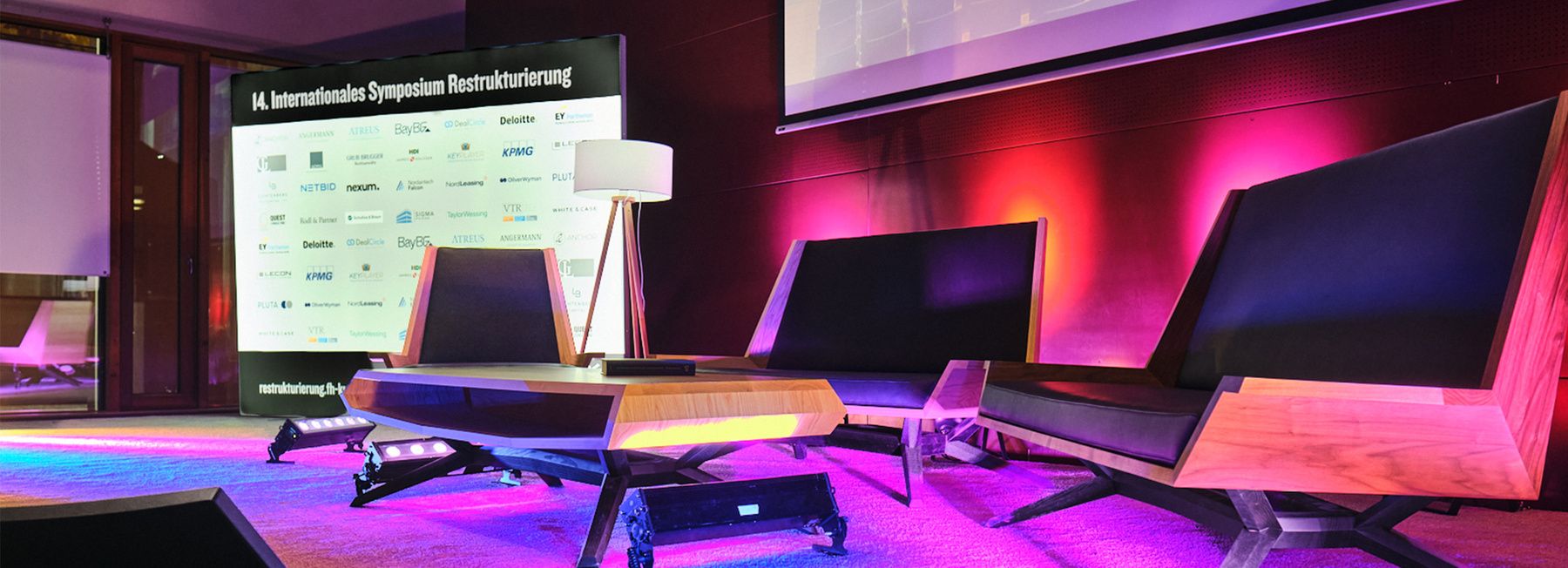 Conference stage at the International Restructuring Symposium featuring modern lounge seating, sponsor backdrop, and purple ambient lighting, prepared for panel discussions at FH Kufstein Tirol.