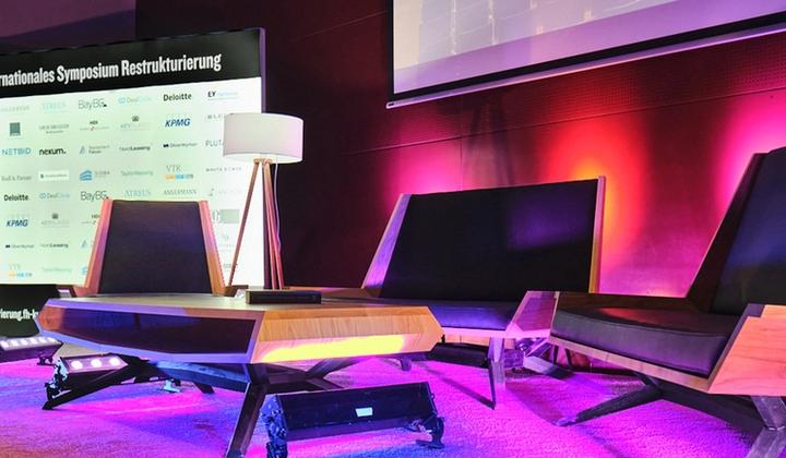 Conference stage at the International Restructuring Symposium featuring modern lounge seating, sponsor backdrop, and purple ambient lighting, prepared for panel discussions at FH Kufstein Tirol.