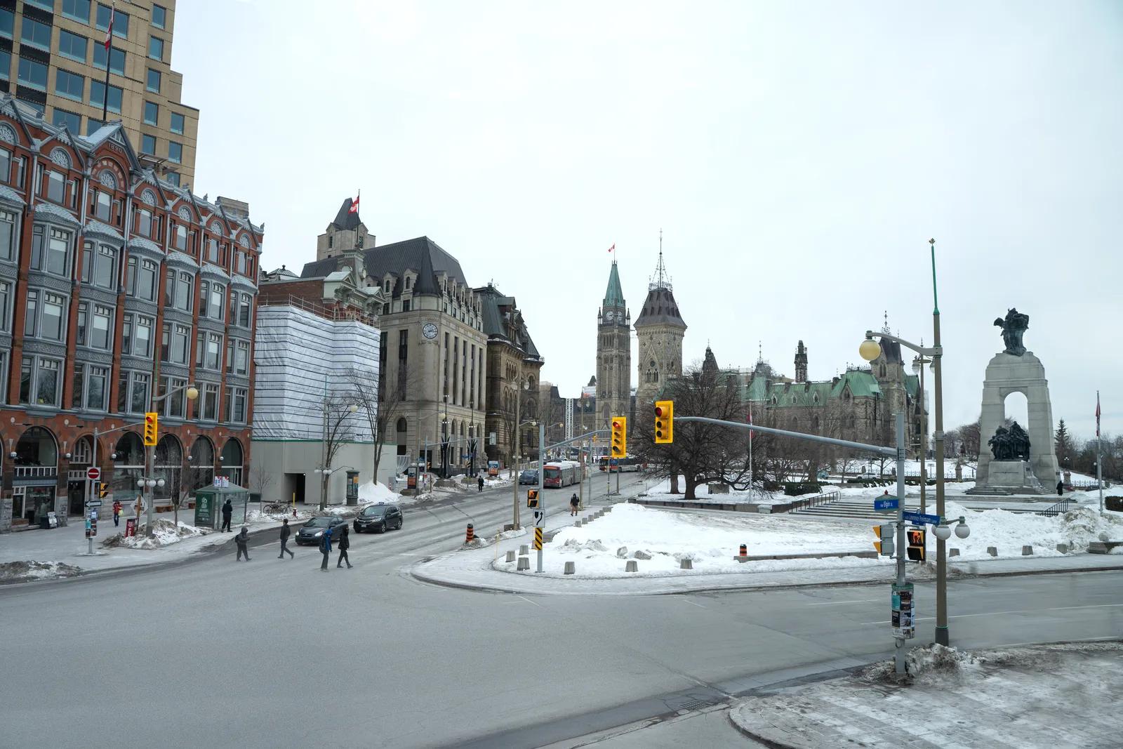 Une vue large, au niveau de la rue, du centre-ville d’Ottawa en hiver, en direction de la tour de la Paix des édifices du Parlement et du Monument commémoratif de guerre du Canada, avec des piétons traversant une intersection bordée de neige.