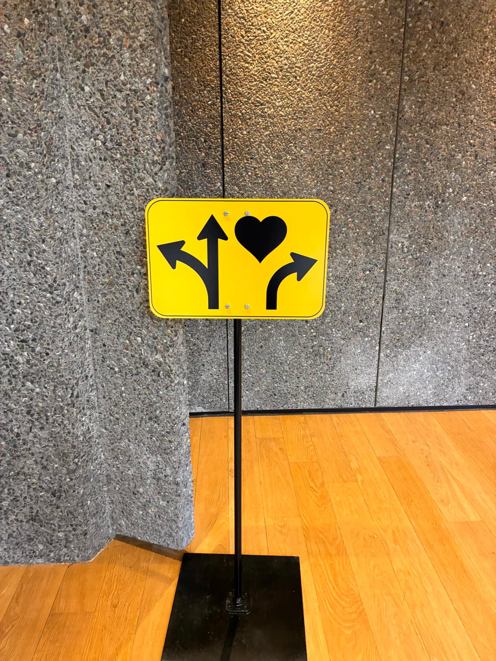 A yellow directional wayfinding sign mounted on a floor stand, showing a forked arrow pointing left and up, a heart, and a curved arrow pointing right, against a textured concrete wall.