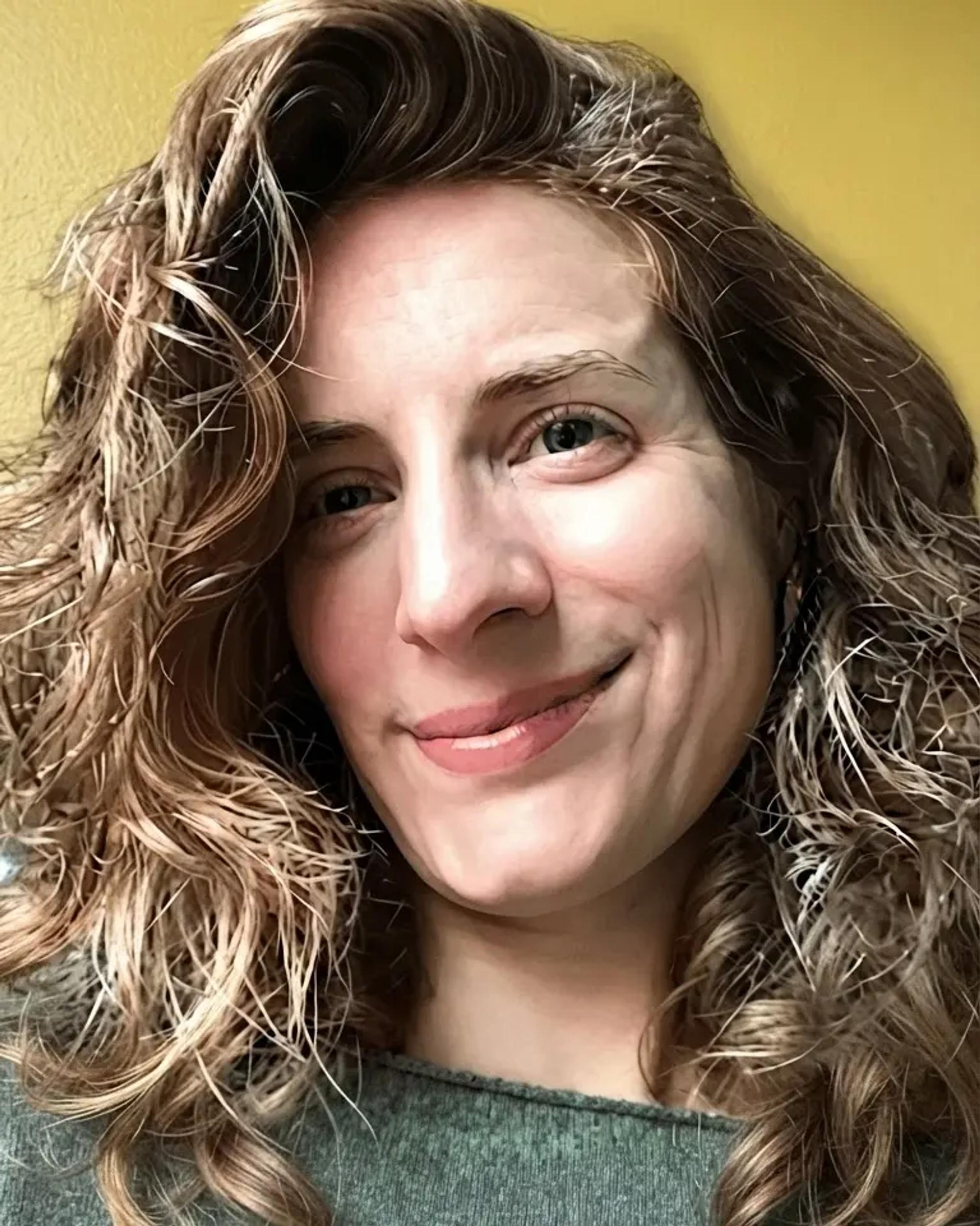 A close-up portrait of Letizia Binda-Partensky smiling warmly, her long wavy brown hair falling loosely around her face, in front of a mustard-yellow wall.