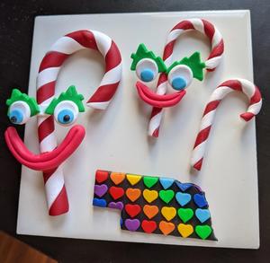 three candy canes made of clay, two with big goofy smiles attached. and one cut out of nebraska that is rainbow hearts on a black background