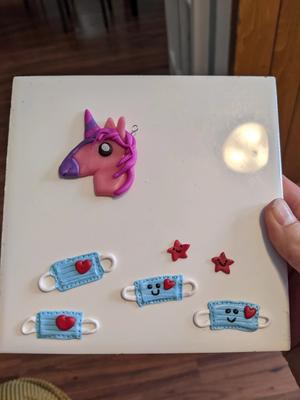 a ceramic tile with a pink clay unicorn and 4 tiny clay face masks with red hearts on them. two of the face masks have tiny smiles