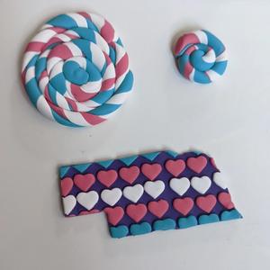 3 items made of polymer clay: two swirly things that are pink, blue, and white, and a Nebraska cutout with hearts in the colors and arrangement of the trans pride flag