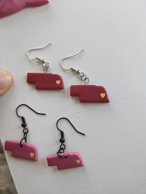 2 pairs of pink Nebraska-shaped earrings, with a yellow heart on the Lincoln spot