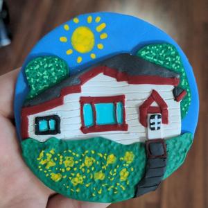 a circle of polymer clay showing a white house with red trim with two green trees in the background