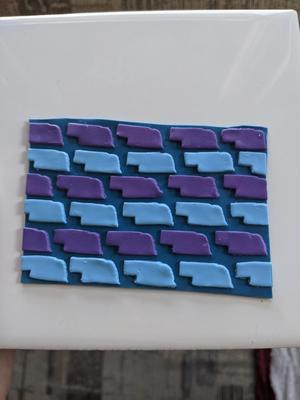 a grid of raised Nebraska shapes on a solid blue background. rows are alternating light blue and purple