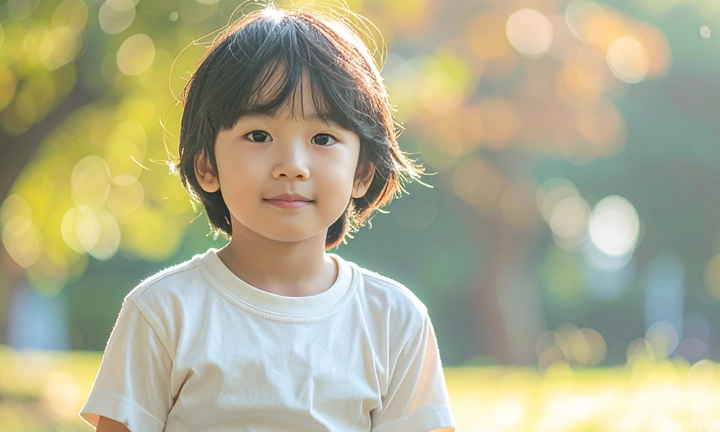 A child in a white T-shirt sits on grass in a park, with blurred trees and bright sunlight behind.