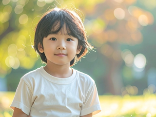 A child in a white T-shirt sits on grass in a park, with blurred trees and bright sunlight behind.