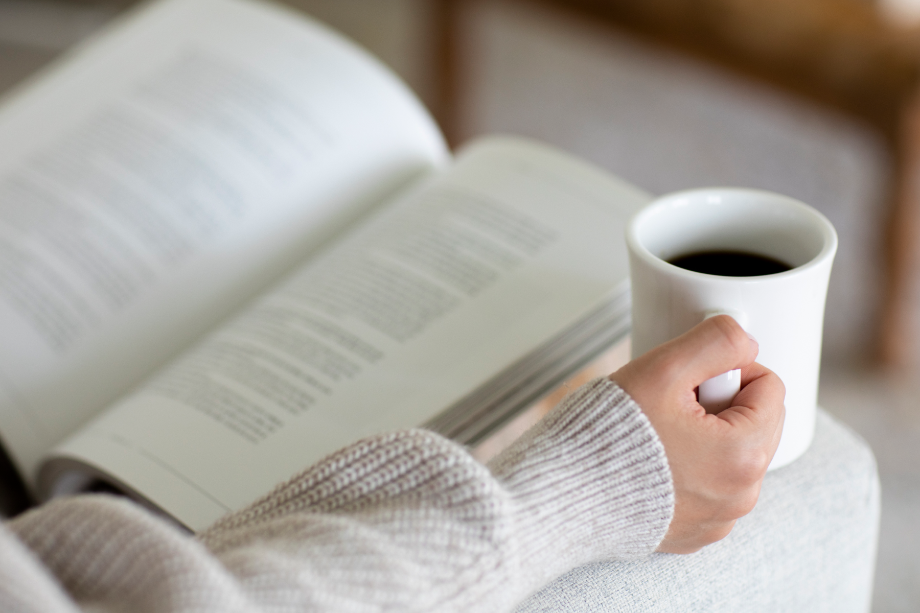 A person holding a coffee cup while reading an open book resting on their lap.