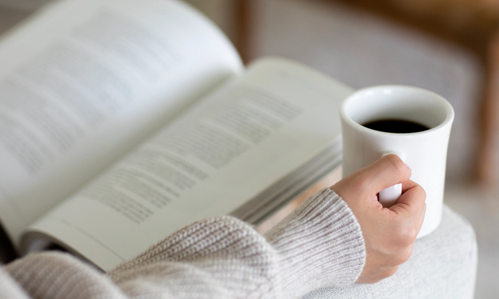 A person holding a coffee cup while reading an open book resting on their lap.
