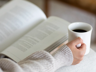 A person holding a coffee cup while reading an open book resting on their lap.