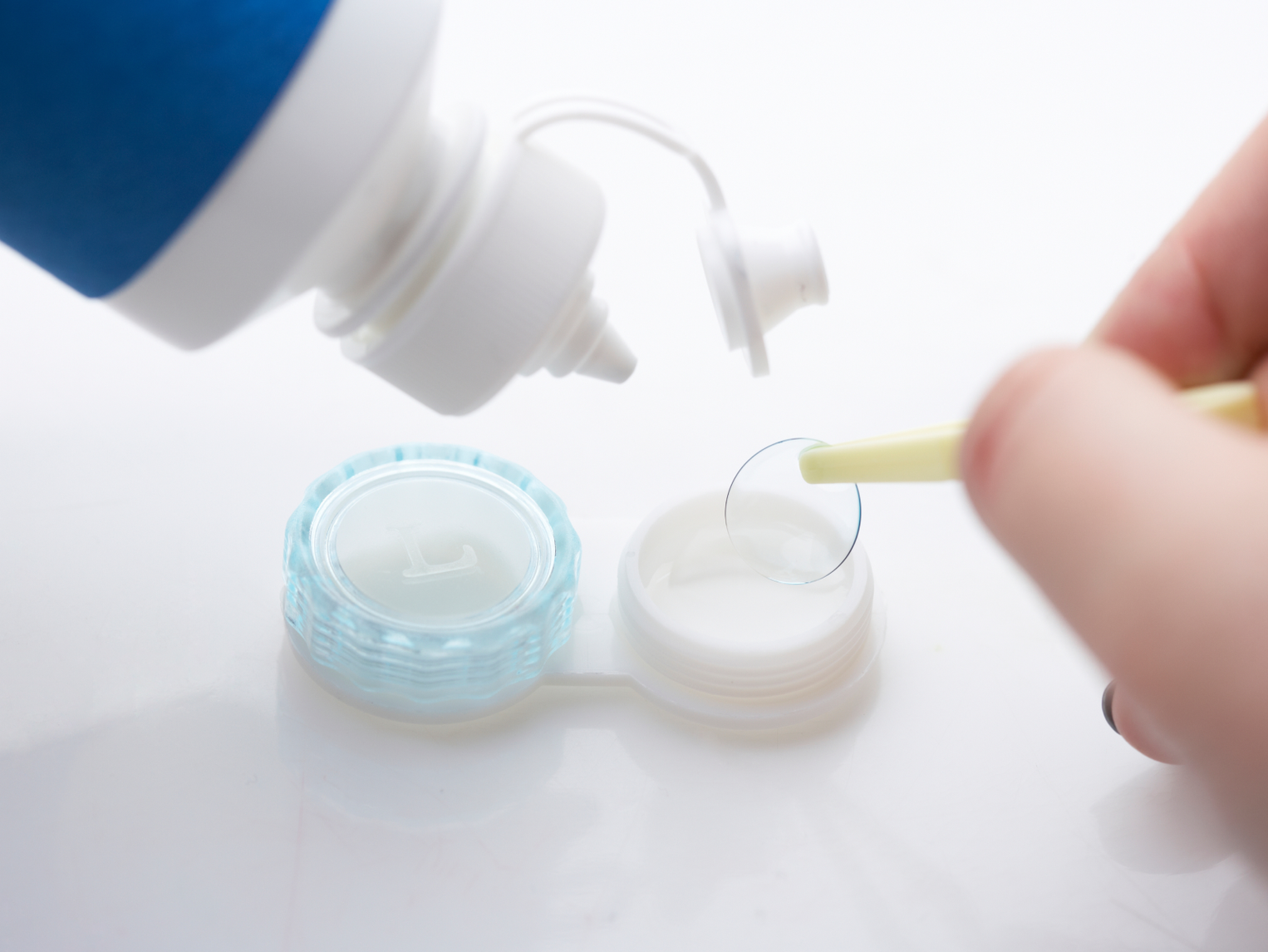 A contact lens held with tweezers above a lens case, with a bottle of solution beside it.