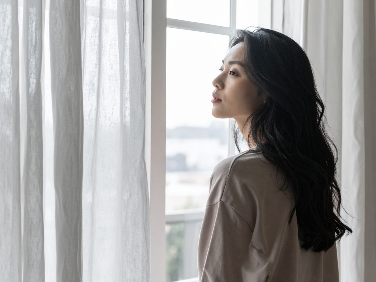 A woman standing beside a window with sheer curtains, looking outside in soft daylight.