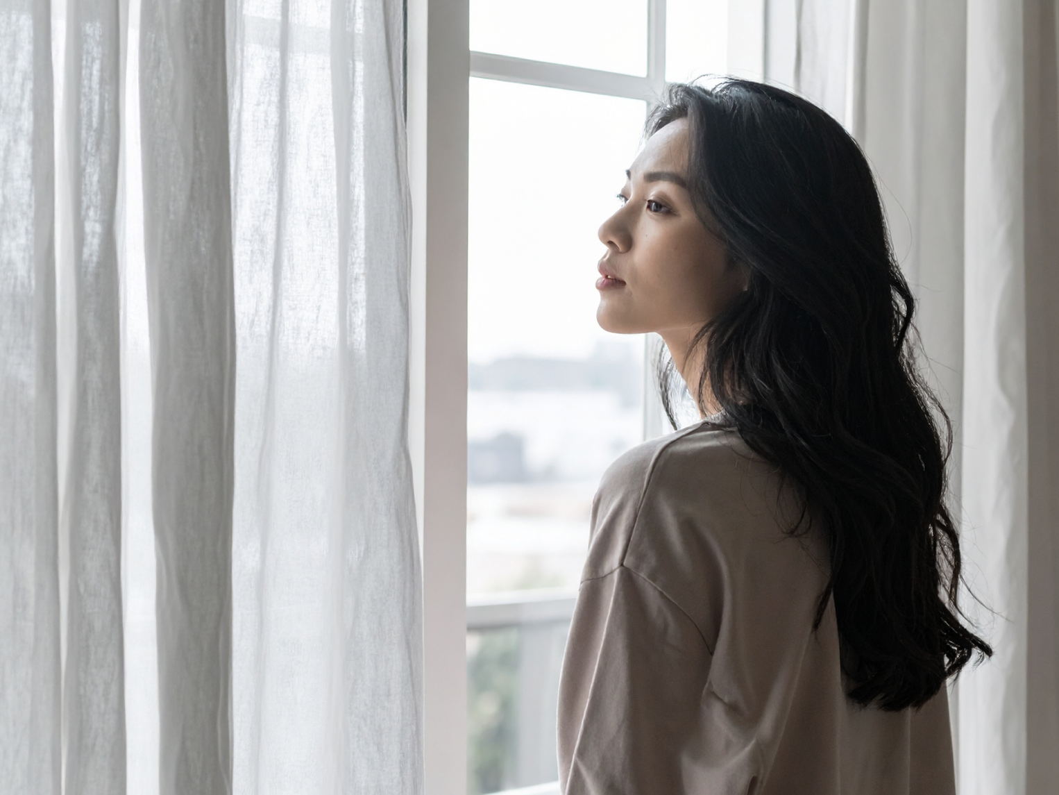 A woman standing beside a window with sheer curtains, looking outside in soft daylight.