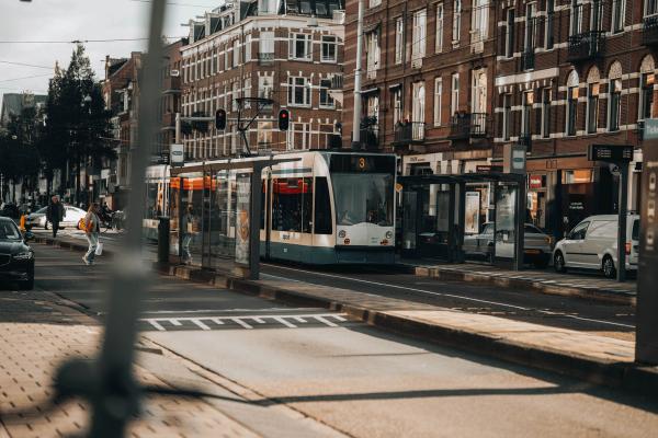 Tram GVB de la ligne 3 arrêté à une station de rue bordée de maisons en brique à Amsterdam, avec un abribus et des passants © Pexels