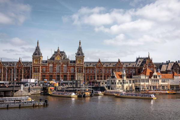La façade en brique de la gare centrale d'Amsterdam vue depuis l'eau, avec les bateaux de croisière amarrés au premier plan © Pexels