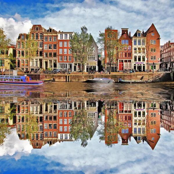 Maisons de canal historiques d'Amsterdam se reflétant parfaitement dans l'eau sous un ciel bleu, avec un bateau de croisière et des barques amarrées © KLM