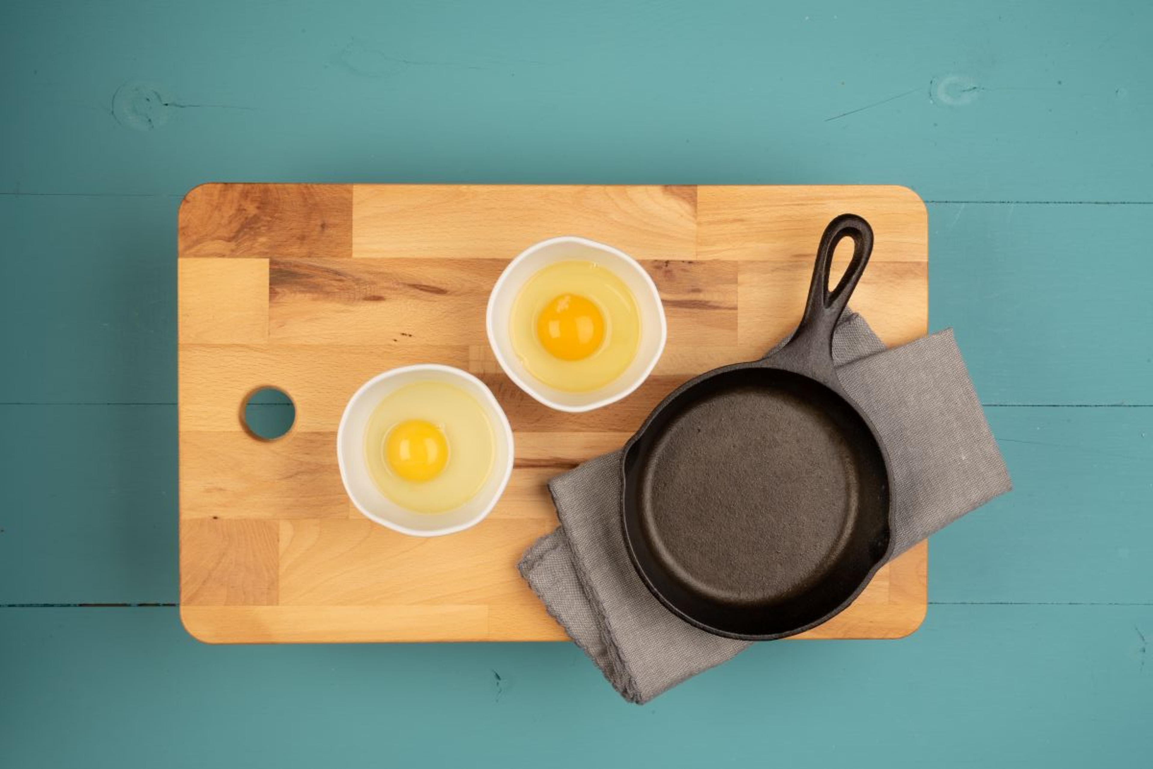 On a cutting board are two raw eggs in separate dishes next to a cast iron pan.