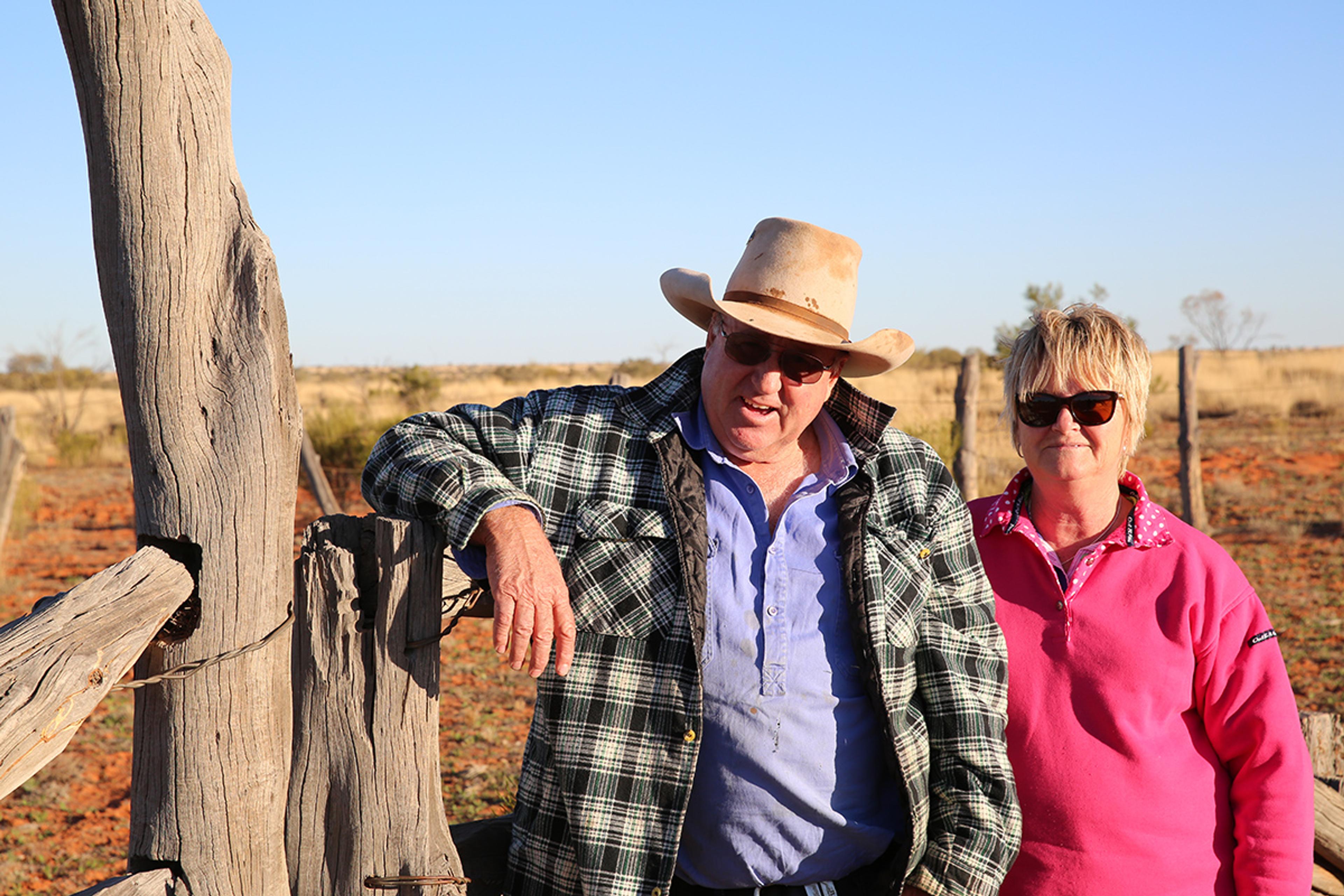 Two Australian grass-fed beef farmers stand in the sun and lean against a wooden post.