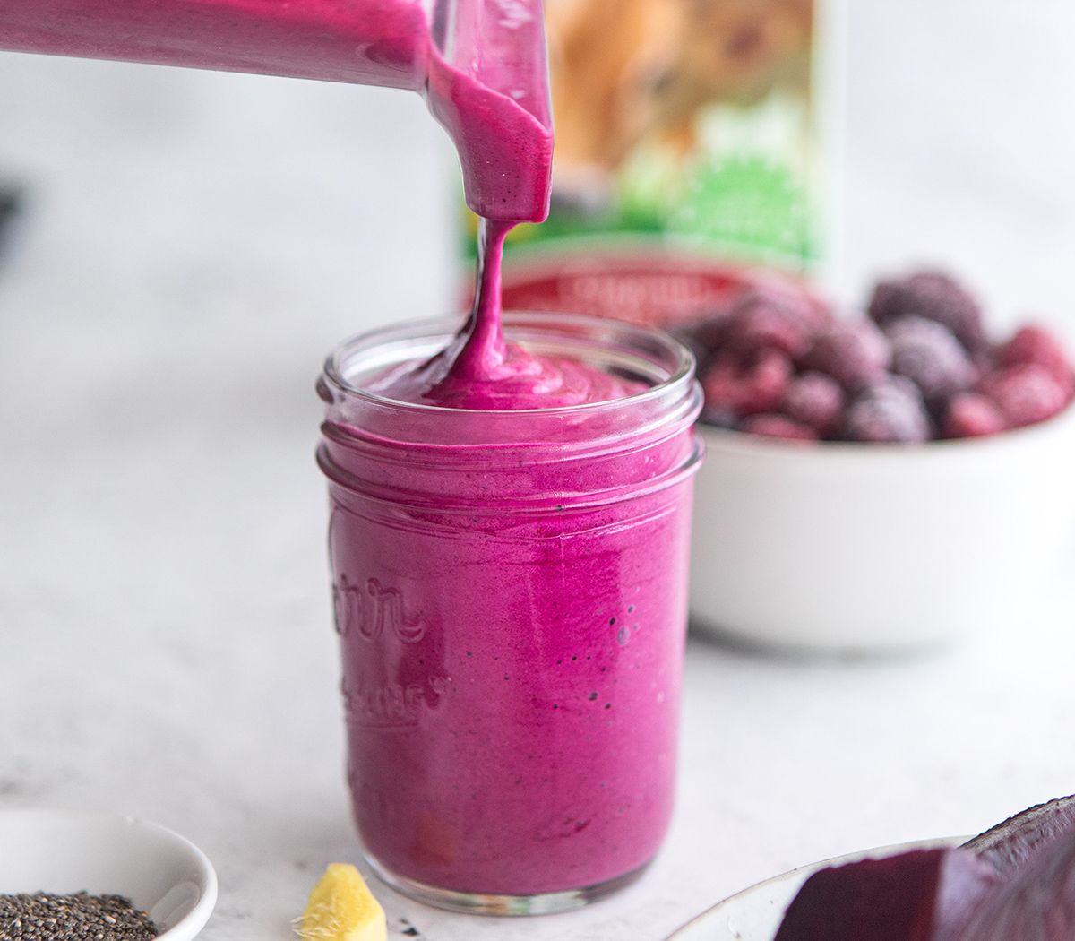 Berry beet smoothie being poured into a mason jar with ginger and chia seeds on the counter.