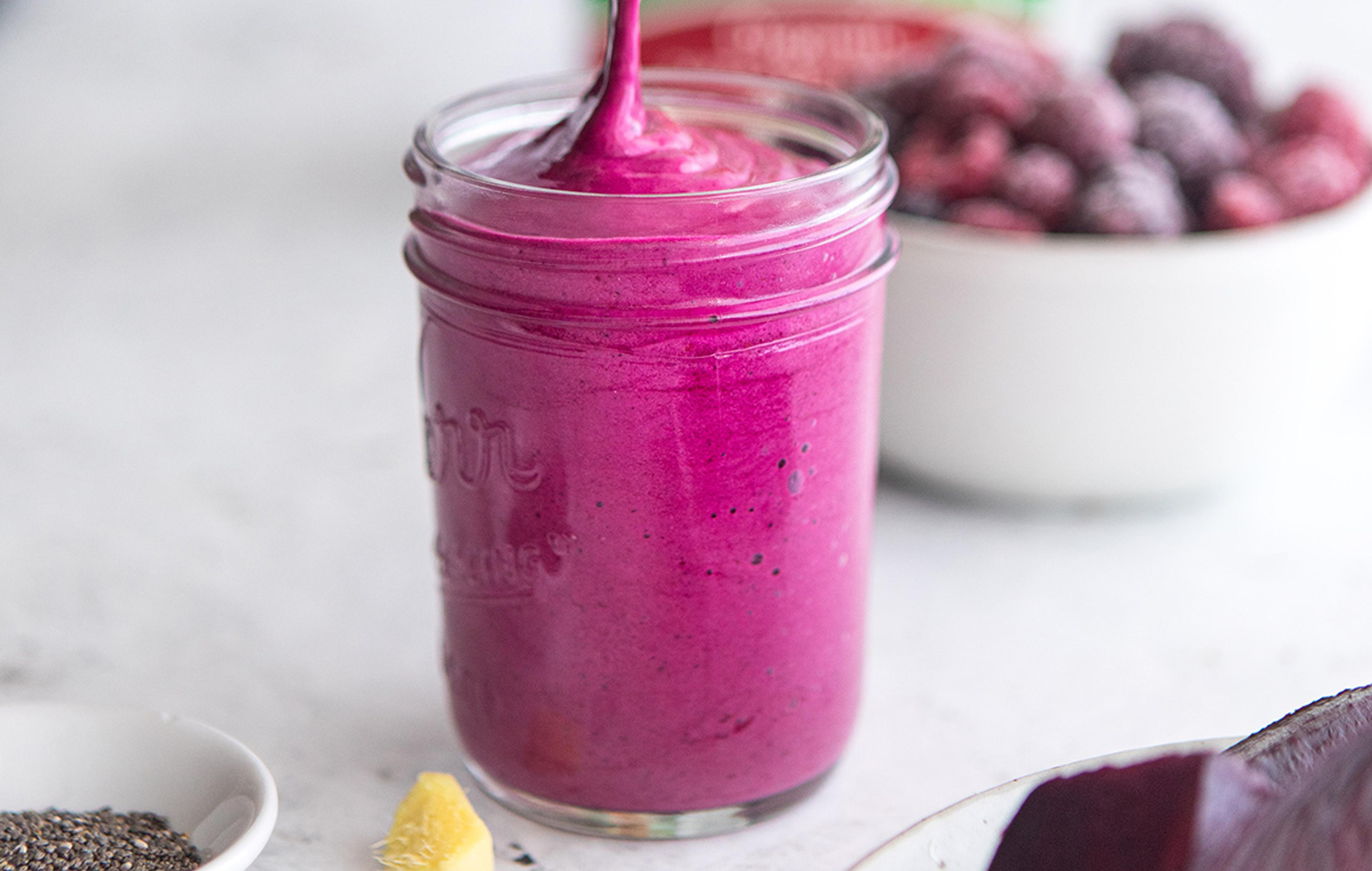 Berry beet smoothie being poured into a mason jar with ginger and chia seeds on the counter.