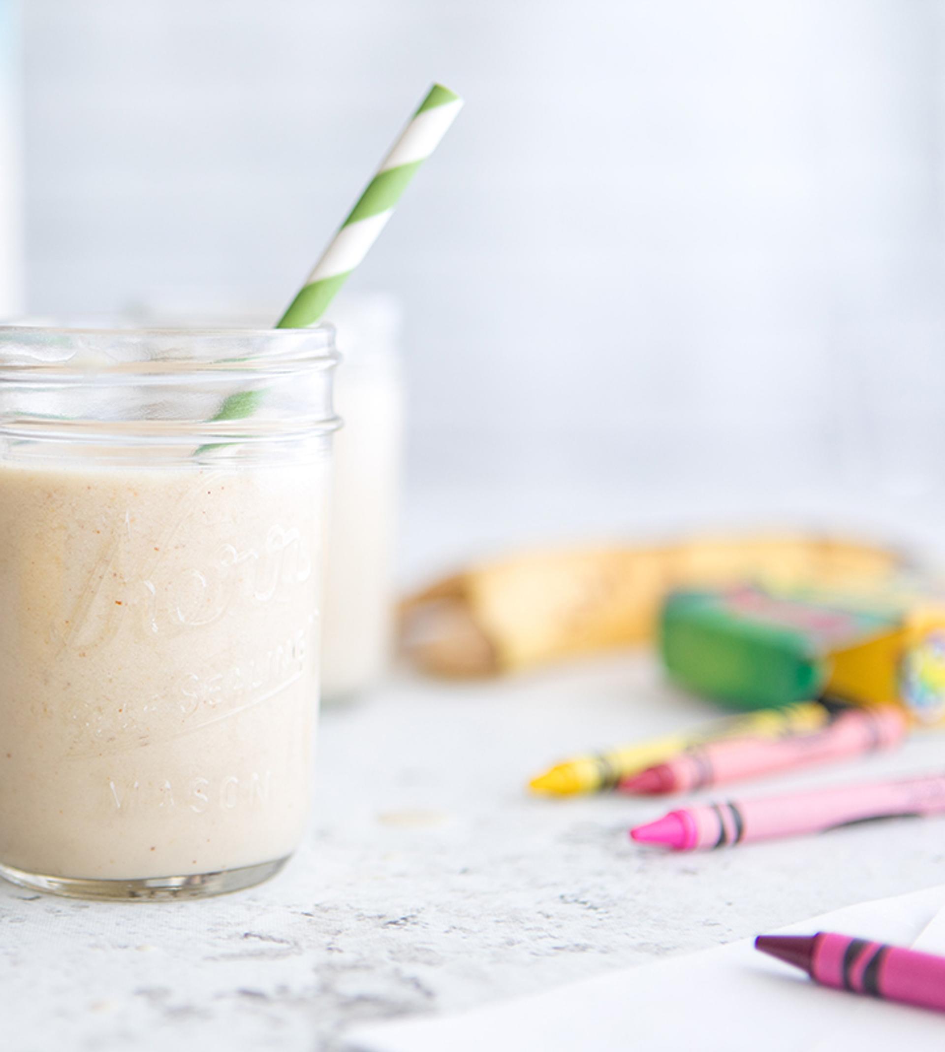 Side view of a light-colored smoothie in a mason jar with a striped green and white straw, and crayons scattered around.