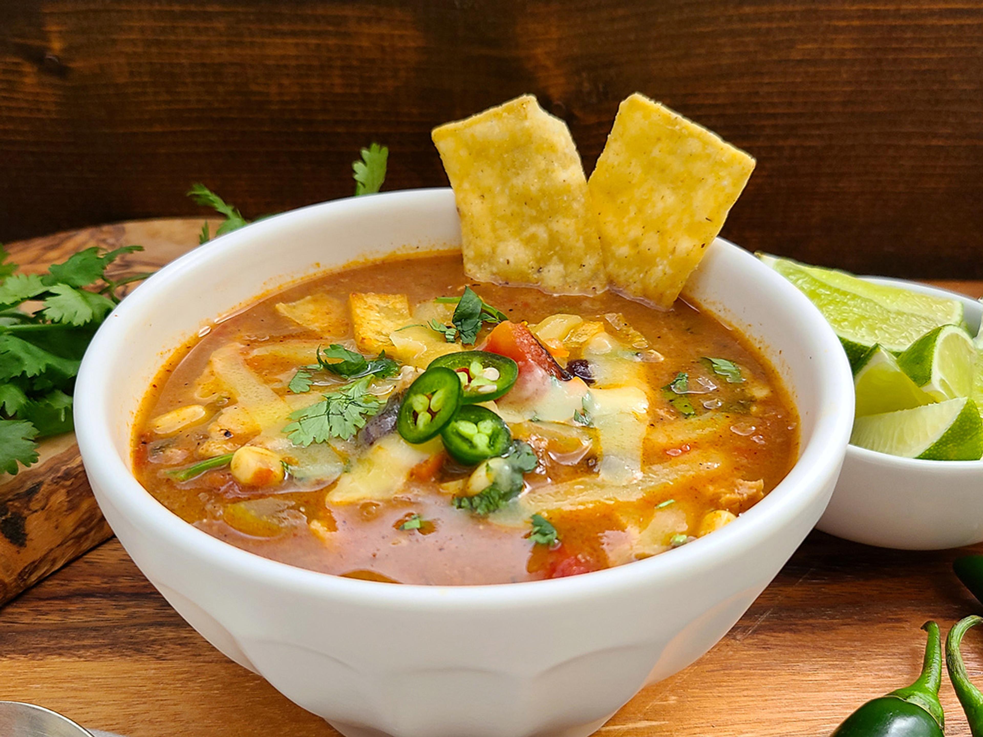 A colorful tomato-based chicken tortilla soup topped with corn chips, shredded cheese, cilantro leaves, and sliced jalapeños.