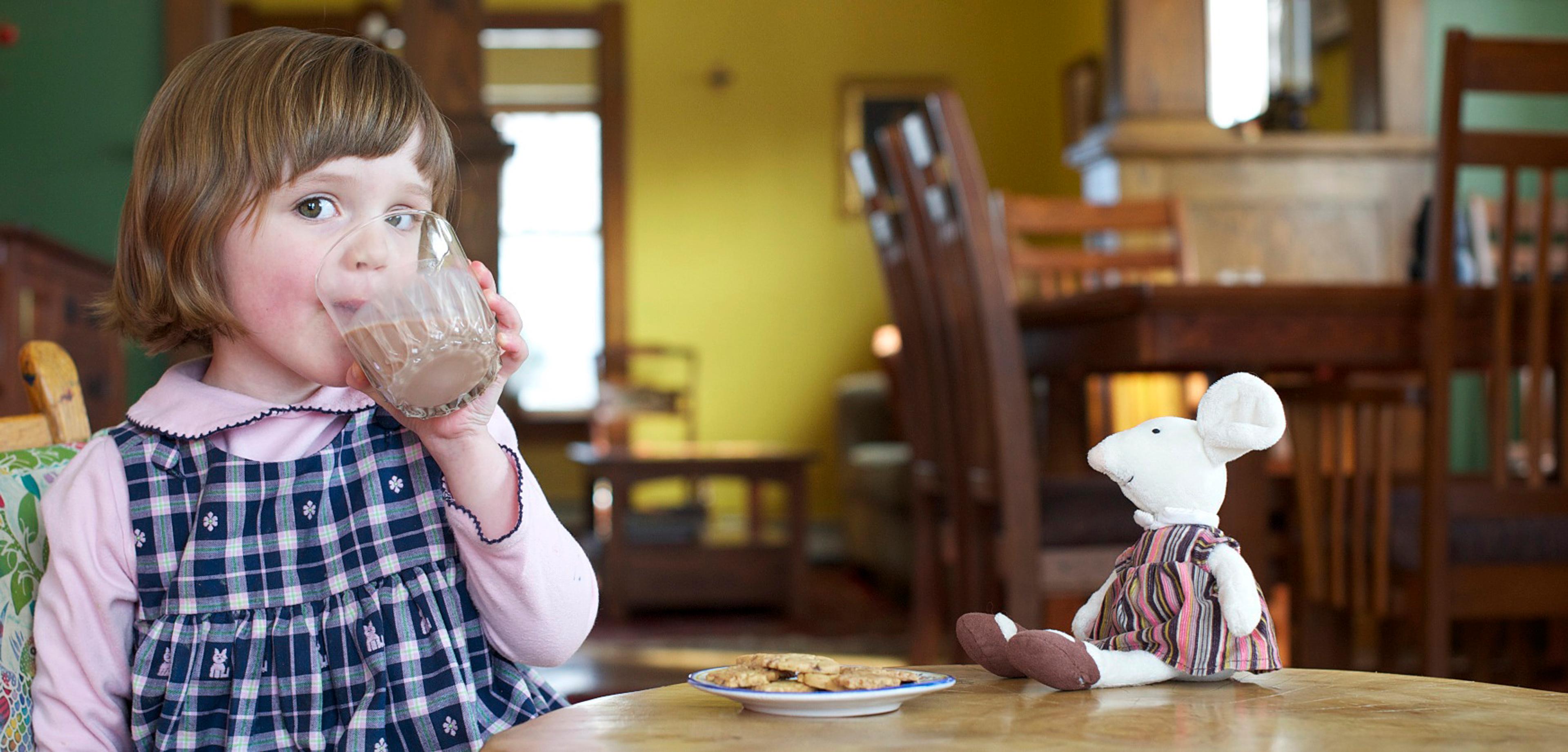A little girl drinks a glass of chocolate milk with her cute toy mouse and a plate of cookies on the table in front of her.
