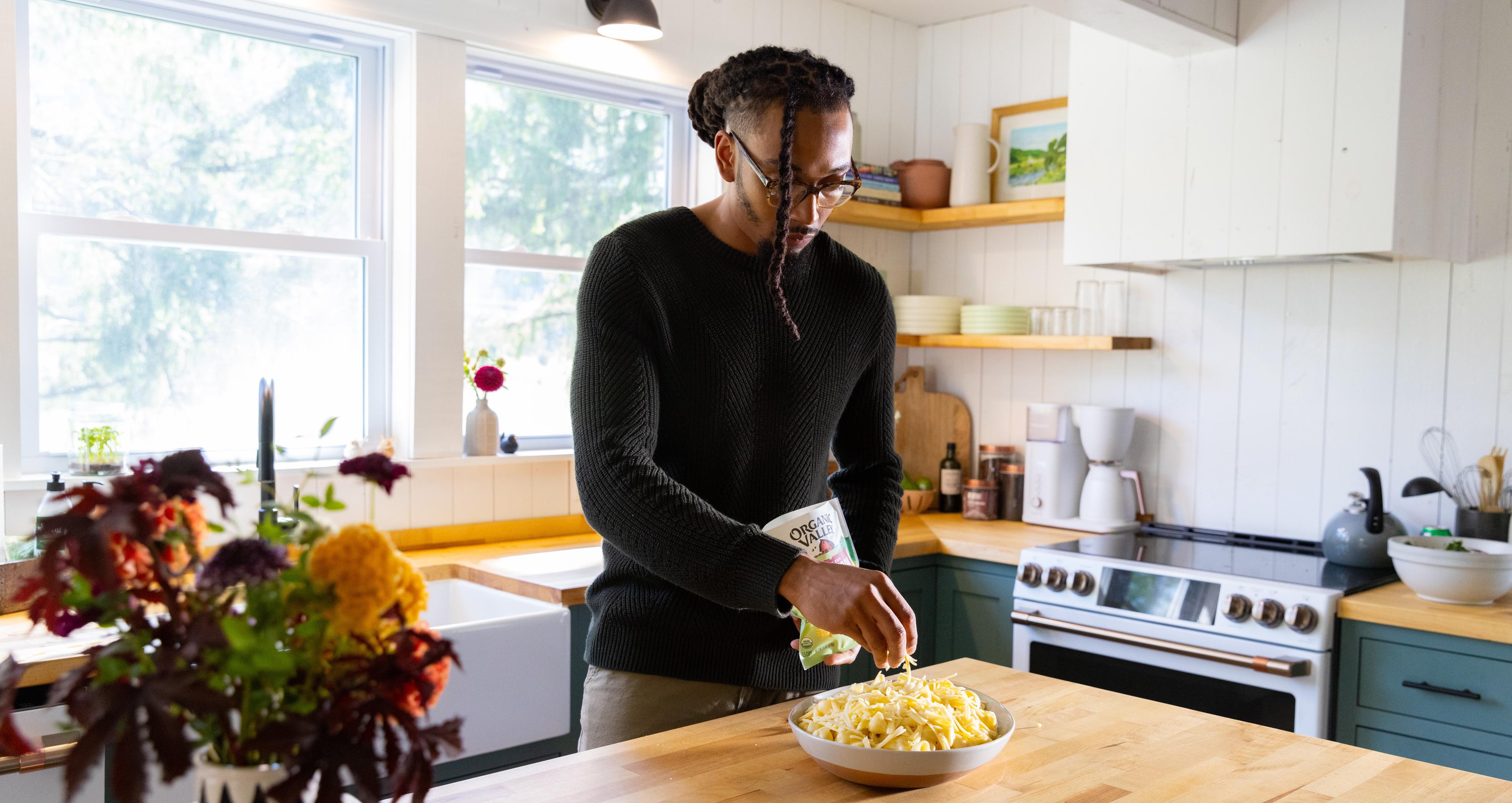 A man sprinkles organic cheddar shredded cheese on a mac and cheese dish.