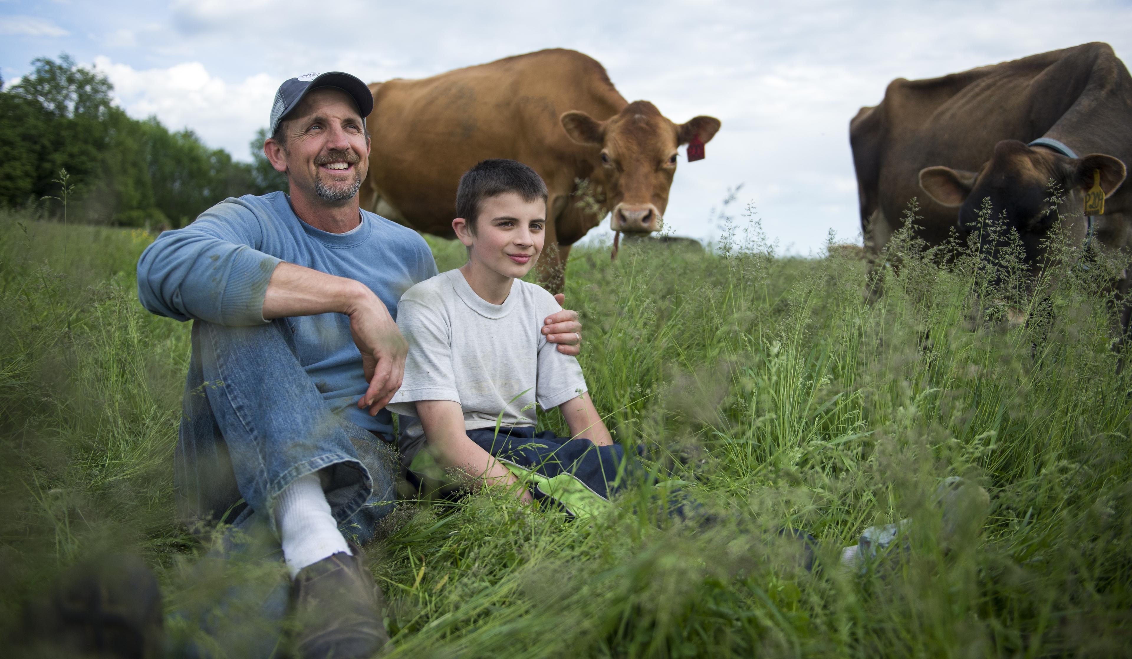 A father and son sit in organic pasture as cows look on.