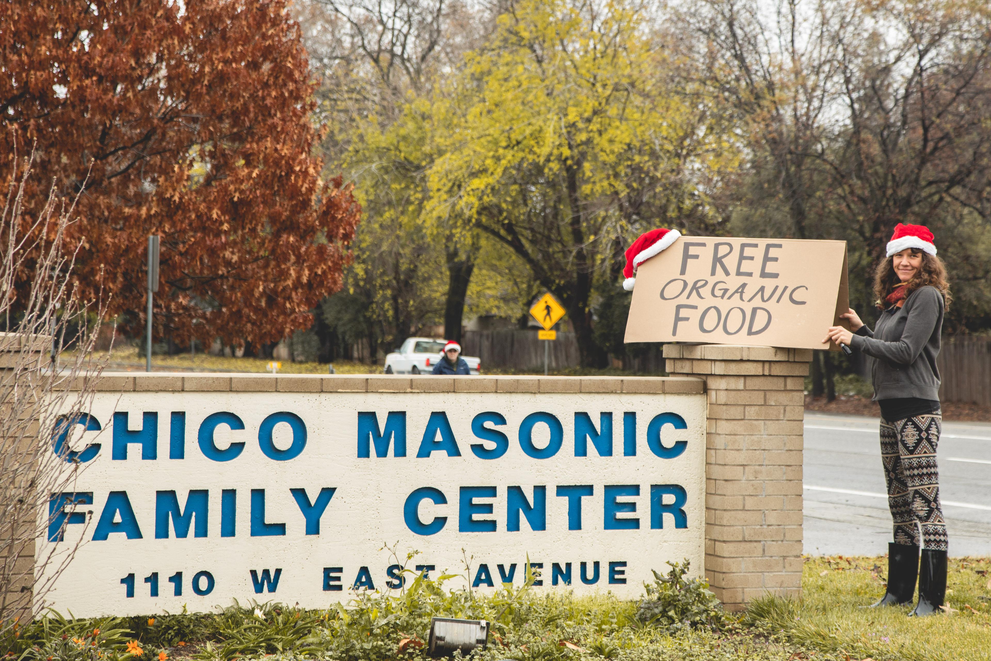 A woman wearing a Santa hat stands beside a sign reading free organic food at the Chico Masonic Family Center.