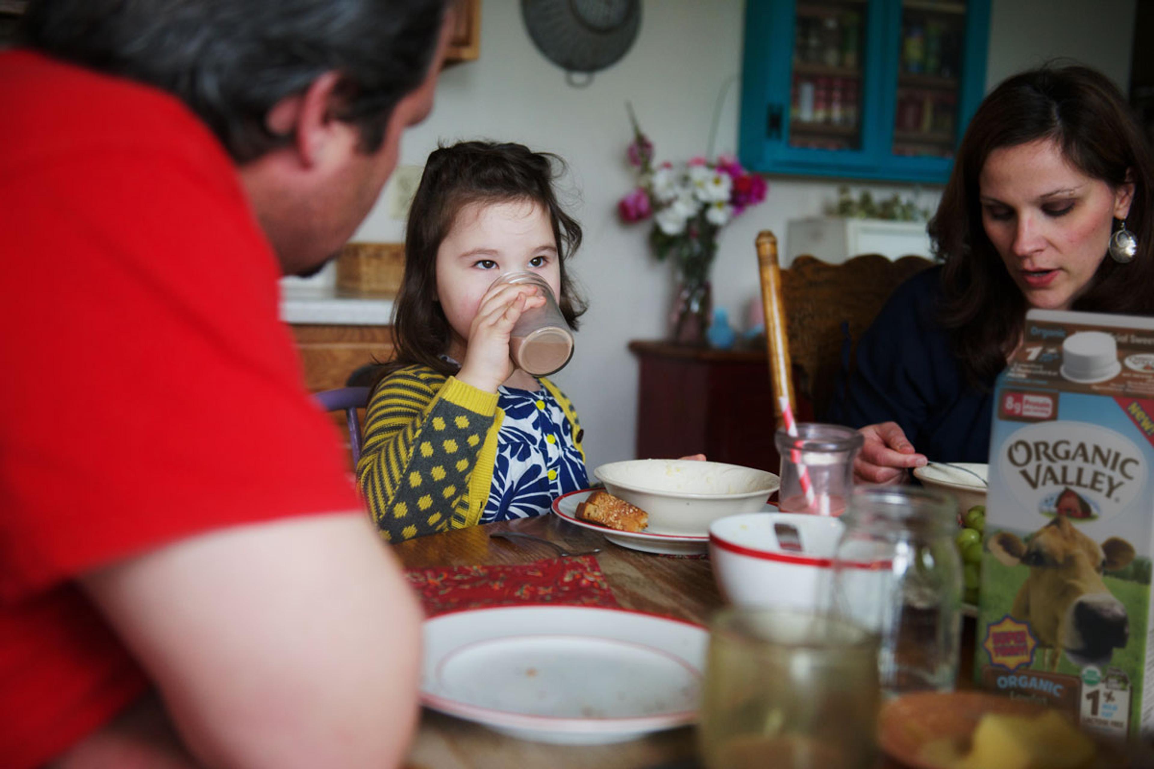 A family is eating at the dinner table, and a little girl is drinking chocolate milk.