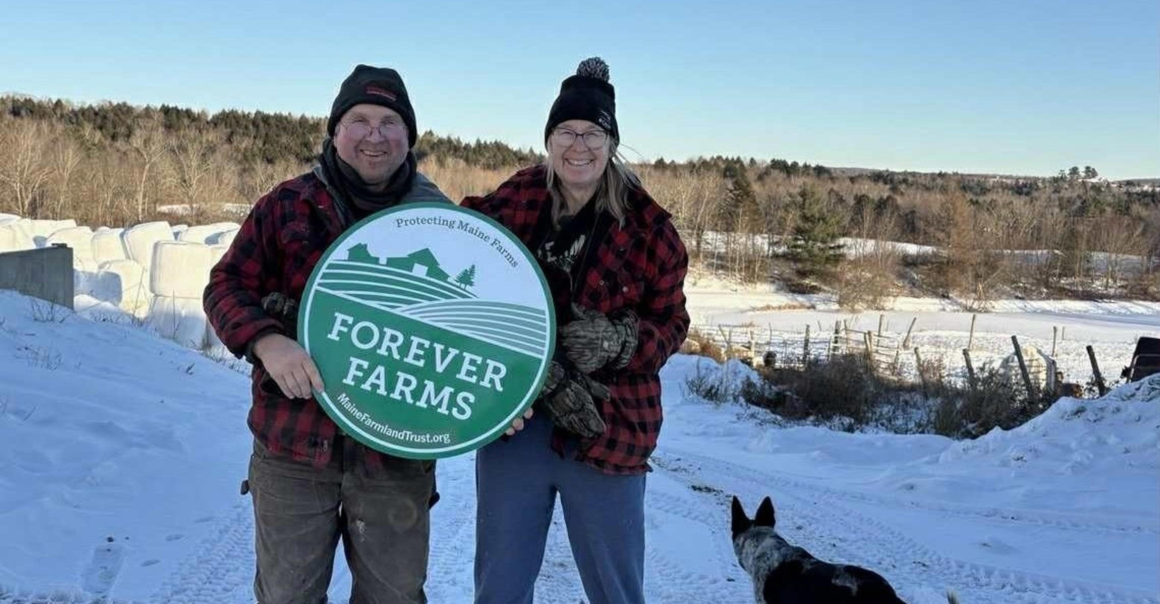 Doug and Linda Hartkopf hold a ‘Forever Farms’ sign as a cattle dog walks along a snowy path.