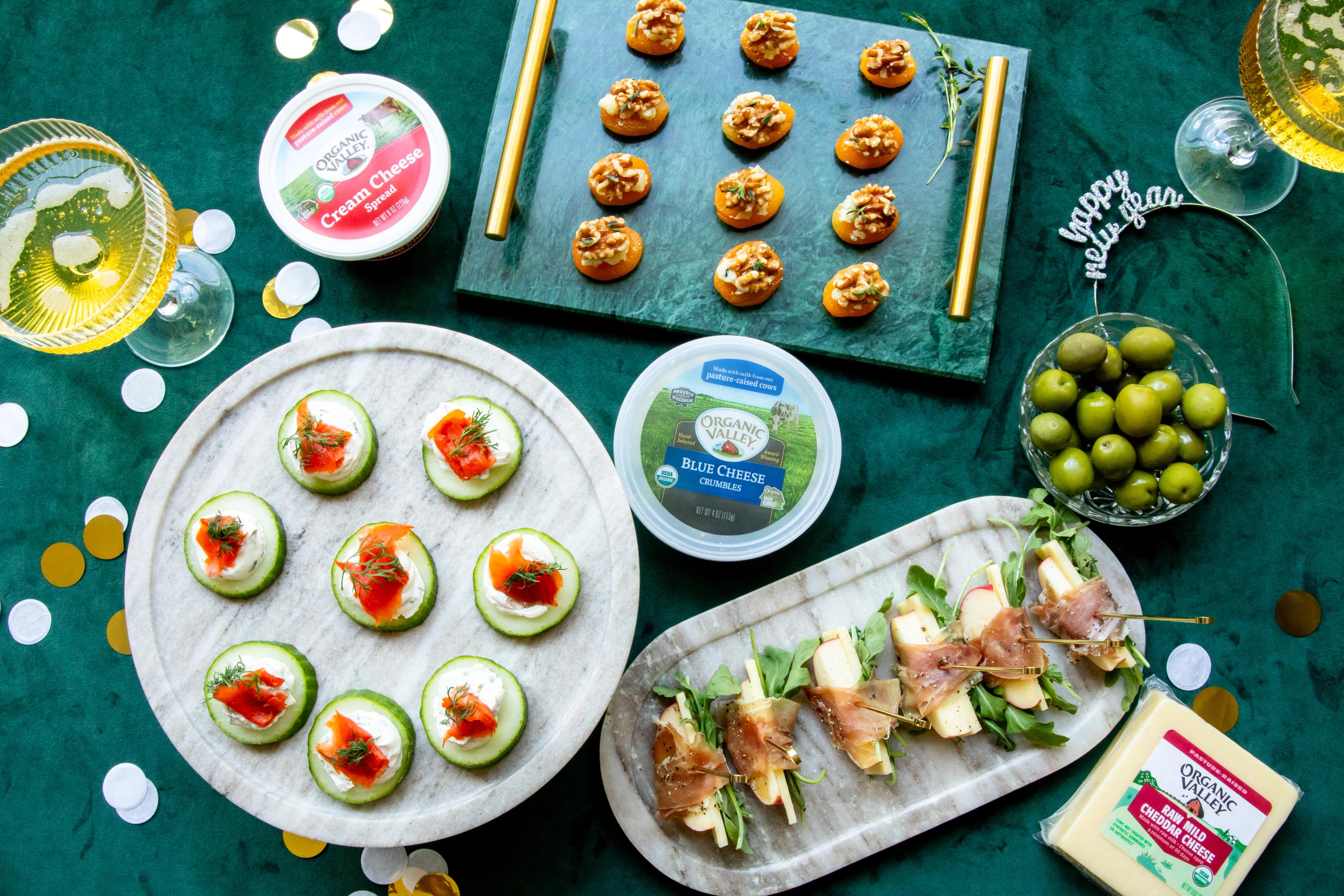 New Year’s Eve finger food, prosciutto bites, stuffed apricots and smoked salmon cucumber snacks on a counter next to packages of organic cheeses, olives and party decor. 