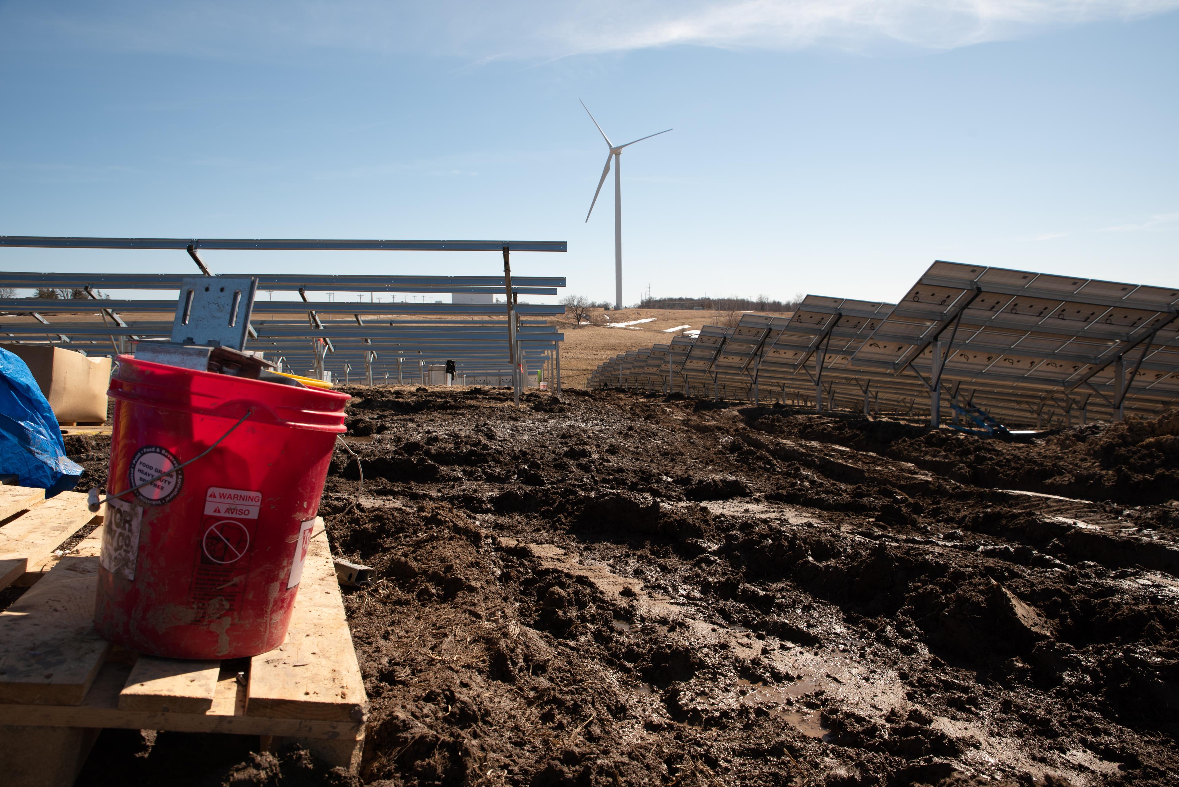 A red bucket on a table overlooking a field of churned, muddy ground, with partially assembled solar panels in rows and a wind turbine in the distance.