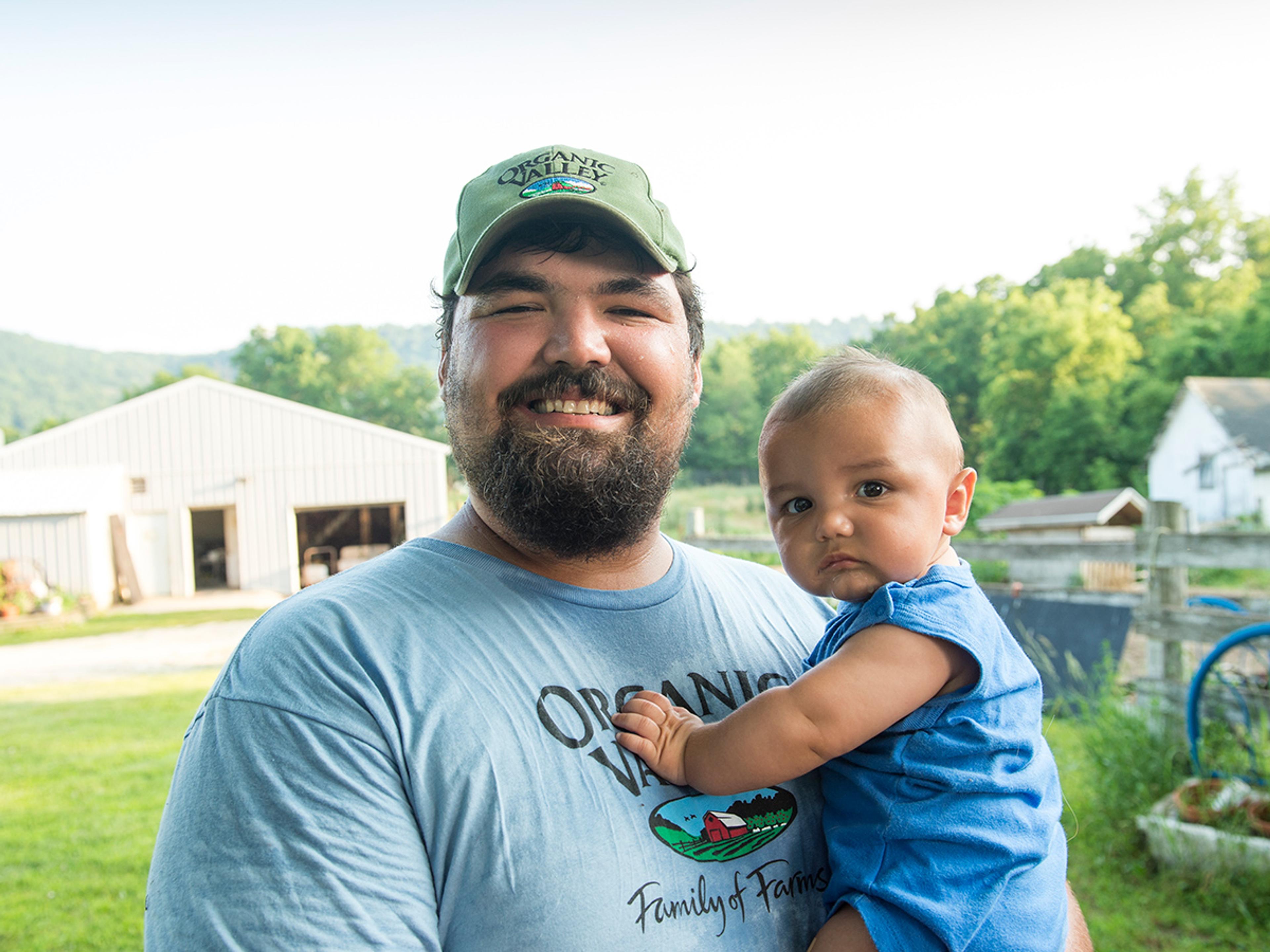 A bearded Joel Goede wearing an Organic Valley baseball cap smiles and holds his baby. 